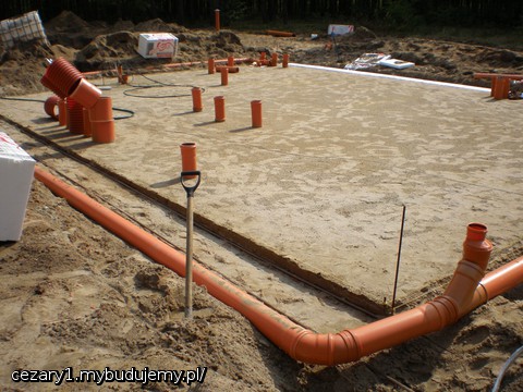 po wkopaniu i ułożeniu przyłączy kanalizacyjnych i podejść pompy ciepła, wody, prądu i ponownym zagęszczeniu podbudowy i jej wyrównaniu poprzez ściągnięcie łatą z 6 metrowego ceownika po wypoziomowanych prętach/szynach umieszczonych wzdłuż płyty pośrodku (1 pręt) i po bokach (2 pręty). Widać również już położone odpływy deszczówki z czterech rynien z rury kanalizacyjnej fi 160mm.